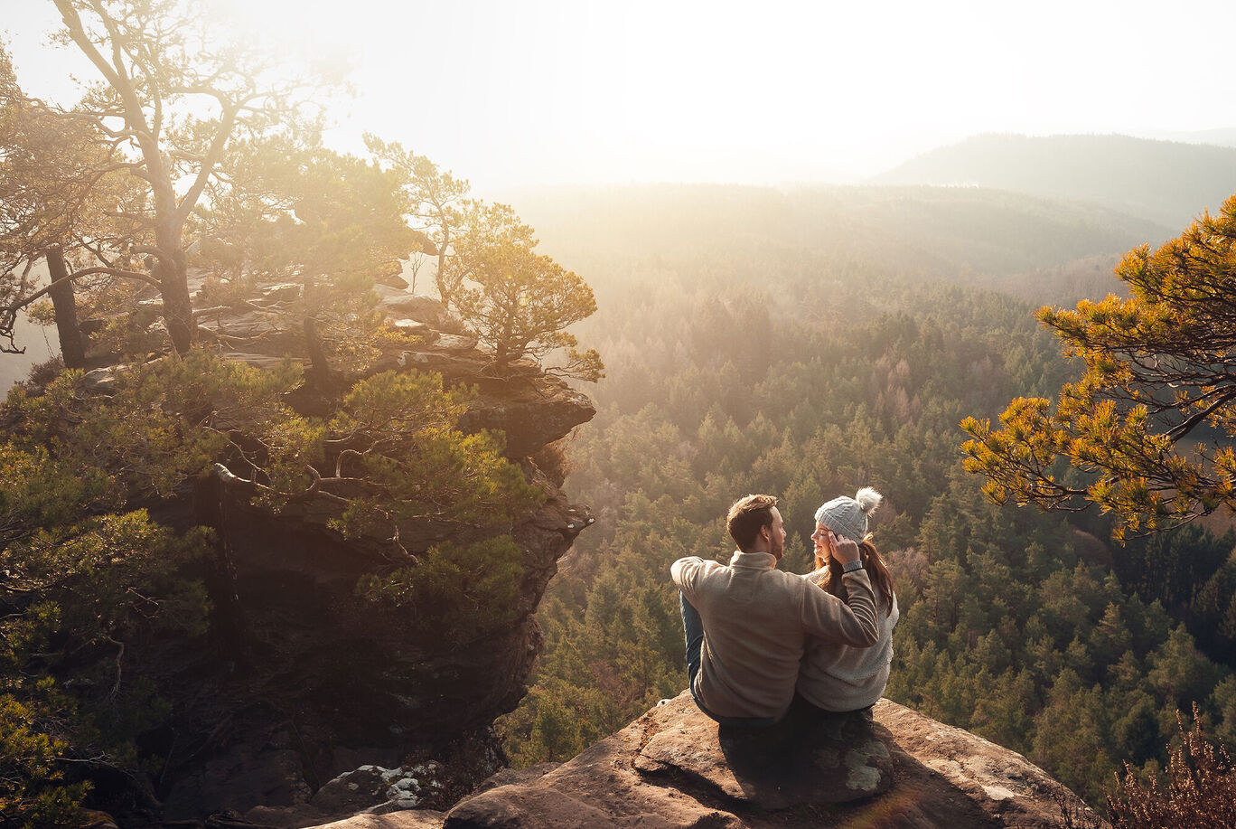Paar sitzt auf einem Felsen und genießt die Aussicht auf die wunderschöne Landschaft mit Wäldern und Hügeln im Sonnenlicht.