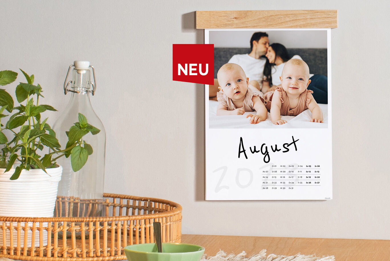 Ein neuer CEWE Fotokalender mit einer Monatsansicht für August, der auf einer Wand hängt. Der Kalender zeigt eine schöne Erinnerung mit einem Baby.