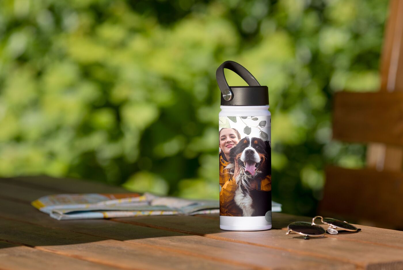 Eine personalisierte Trinkflasche steht auf einem Holztisch und zeigt ein Bild eines Hundes. Im Hintergrund sind grüne Pflanzen zu sehen.