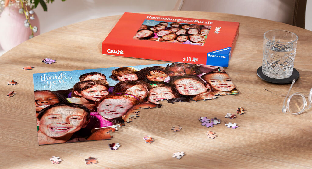 Puzzle mit fröhlichem Gruppenbild von vielen lächelnden Personen, daneben die Verpackung des cewe Revue-Puzzles.