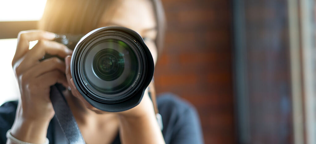Eine Person hält eine Kamera und fotografiert. Der Hintergrund zeigt eine kreative Umgebung mit warmem Licht.