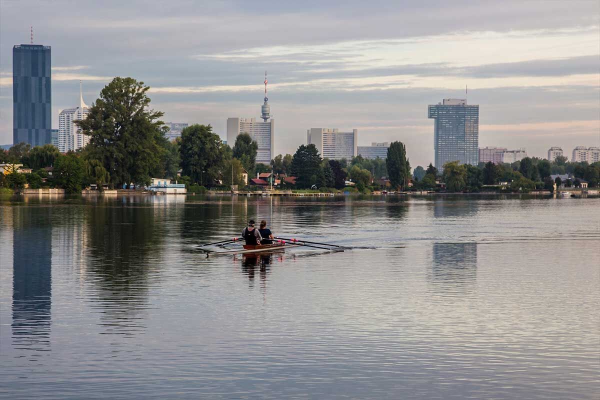 Zwei Personen rudern in einem Boot auf einem ruhigen See, umgeben von Gebäuden und grünen Bäumen