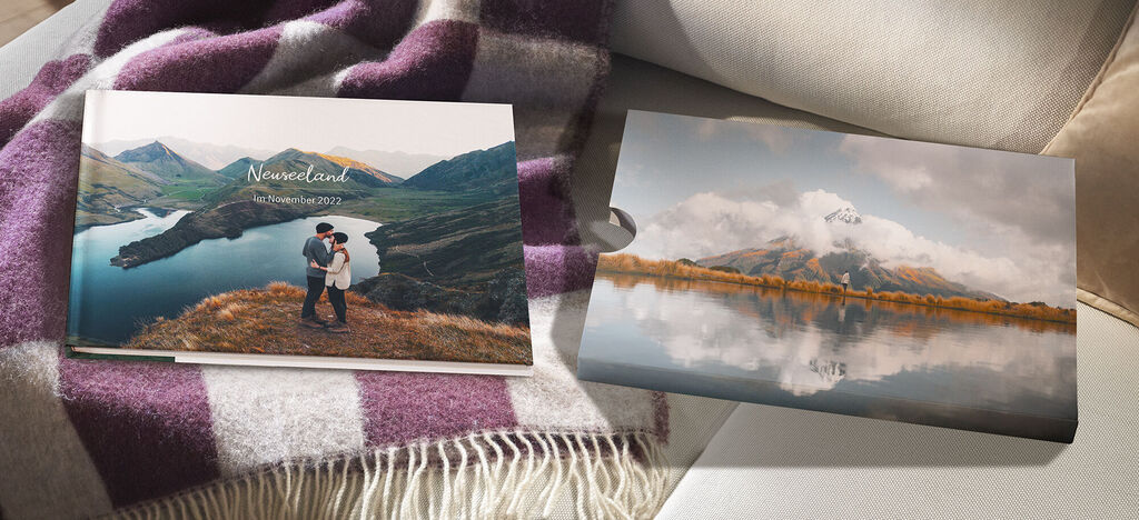 Zwei Fotobücher vor einem gemütlichen Hintergrund, eines zeigt eine Liebesaufnahme in der Natur und das andere eine malerische Berglandschaft.
