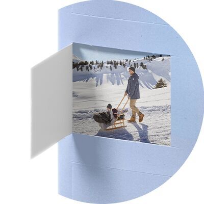 Ein Mann zieht einen Freund auf einem Schlitten durch den Schnee. Im Hintergrund sieht man verschneite Berge unter blauem Himmel. Es ist ein fröhlicher Wintertag.