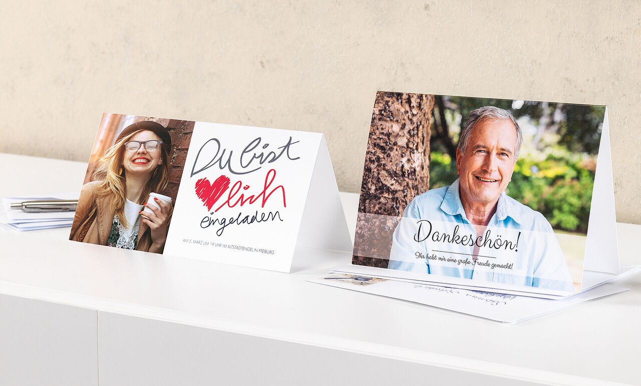 Zwei beschriftete CEWE Grußkarten auf einem Tisch, links eine Karte mit einem Herzen und Text, rechts eine Dankeskarte mit einer freundlichen Person.