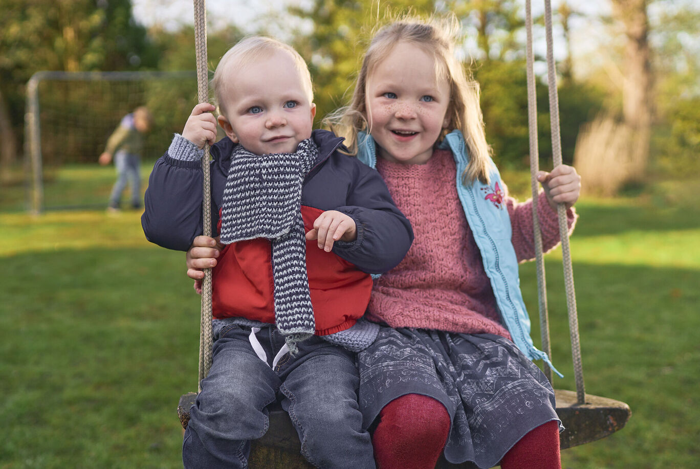 Zwei Kinder sitzen lächelnd auf einer Schaukel im Freien.