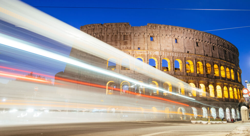 Beleuchtetes Colosseum vor klarem Nachthimmel. Im Vordergrund sind Lichtstreifen von vorbeifahrenden Fahrzeugen zu sehen.
