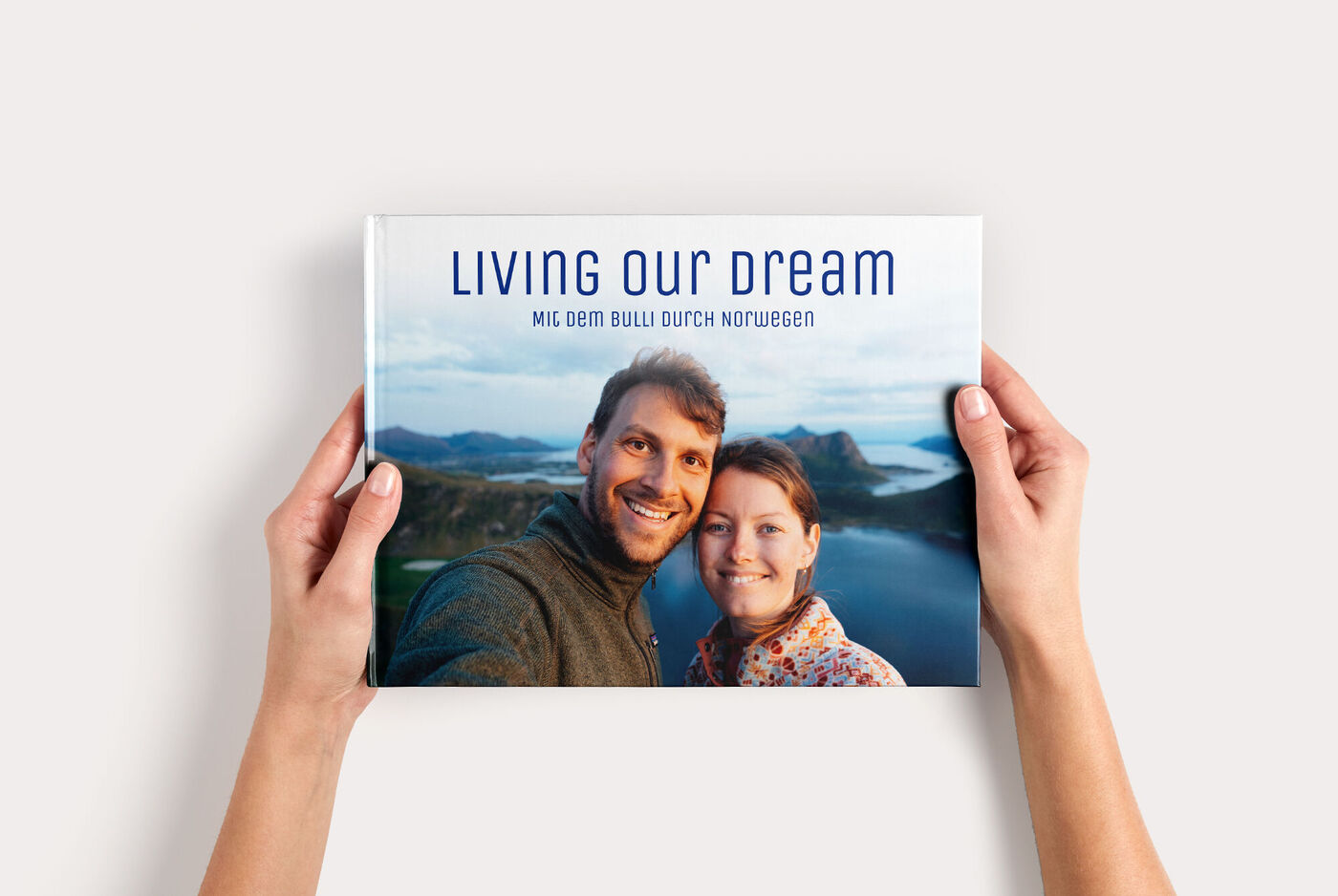 Zwei Hände halten ein CEWE FOTOBUCH Groß Panorama mit einem Paarfoto vor einem Fjord und dem Titel "LIVING OUR DREAM"