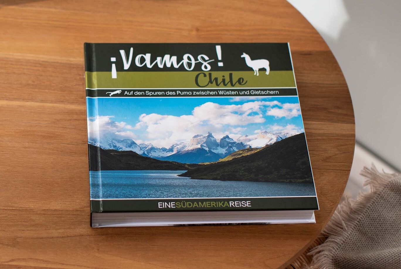 Ein CEWE FOTOBUCH mit dem Titel ¡Vamos! Chile auf einem Holztisch. Das Buch zeigt Bilder von Bergen und Seen in Chile.