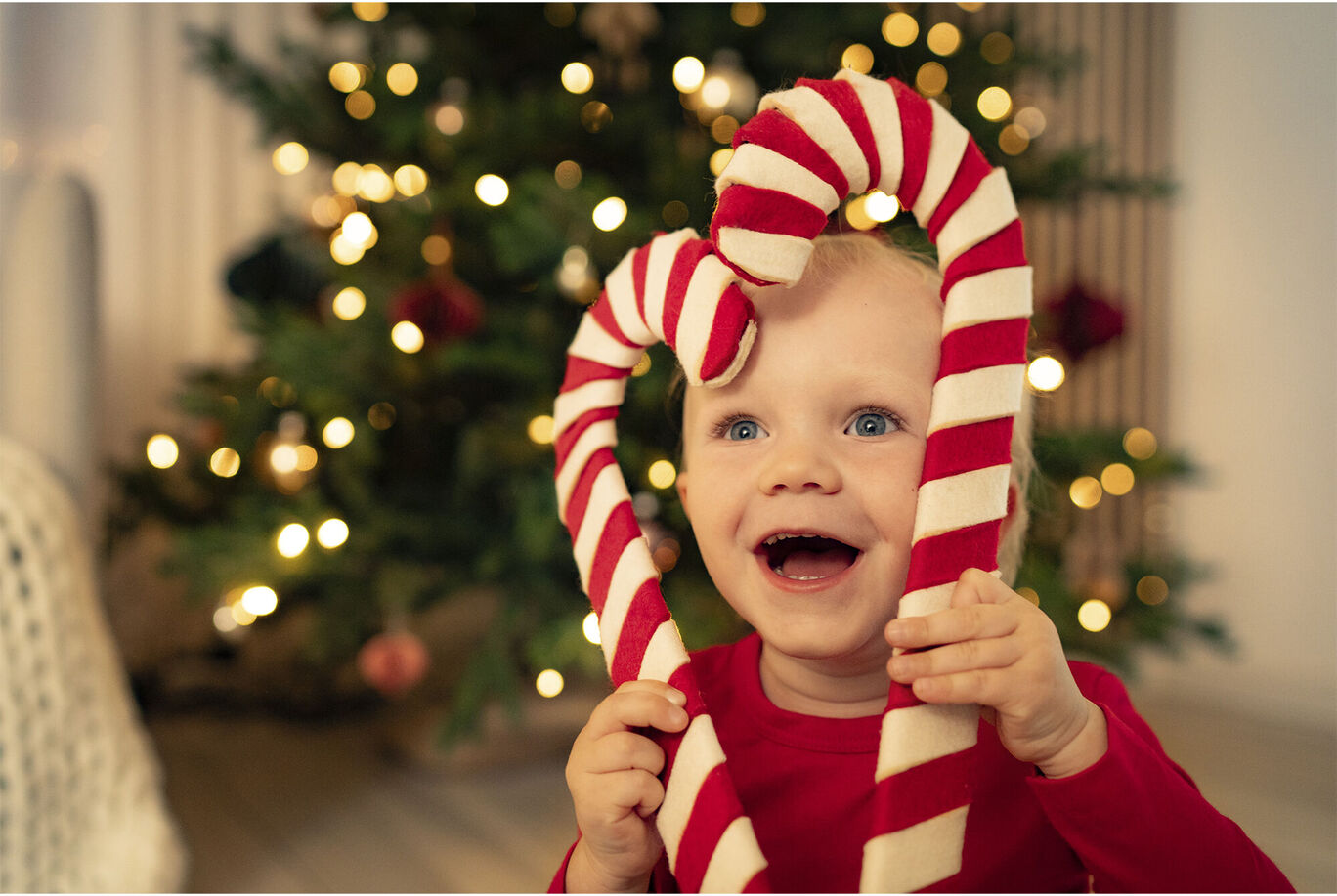 Ein Kind hält zwei rot-weiße Zuckerstangen in Herzform vor einem festlich geschmückten Weihnachtsbaum