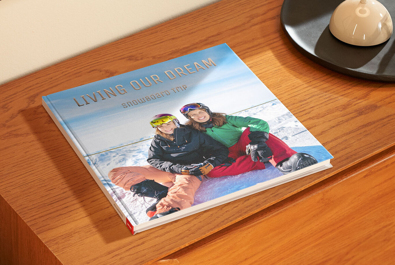 Ein CEWE FOTOBUCH auf einem Tisch. Titel: 'Living Our Dream'. Ein Paar sitzt glücklich im Schnee und genießt ihren Skiurlaub.