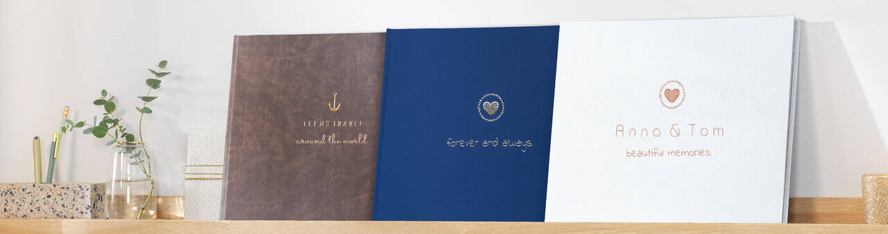 Drei wunderschöne Fotobücher von CEWE stehen auf einem Regal. Verschiedene Designs in Braun, Blau und Weiß.