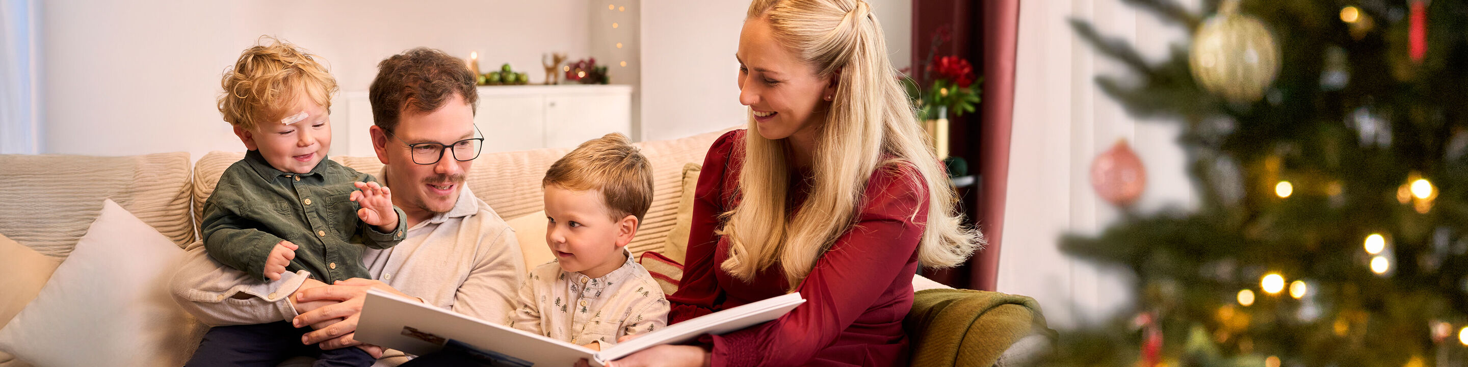 Eine Familie mit zwei kleinen Kinder auf einem Sofa schaut gemeinsam in ein CEWE FOTOBUCH in weihnachtlicher Atmosphäre.