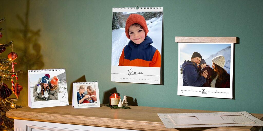 Zwei schöne Fotokalender im Wohnraum, einer mit einem wintersportlichen Motiv und einer mit einem liebevollen Familienbild, Beiges auf vertiefter Holzfläche.