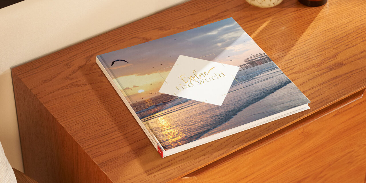 Ein CEWE FOTOBUCH liegt auf einem Holzsideboard. Es zeigt einen Sonnenuntergang über dem Wasser.