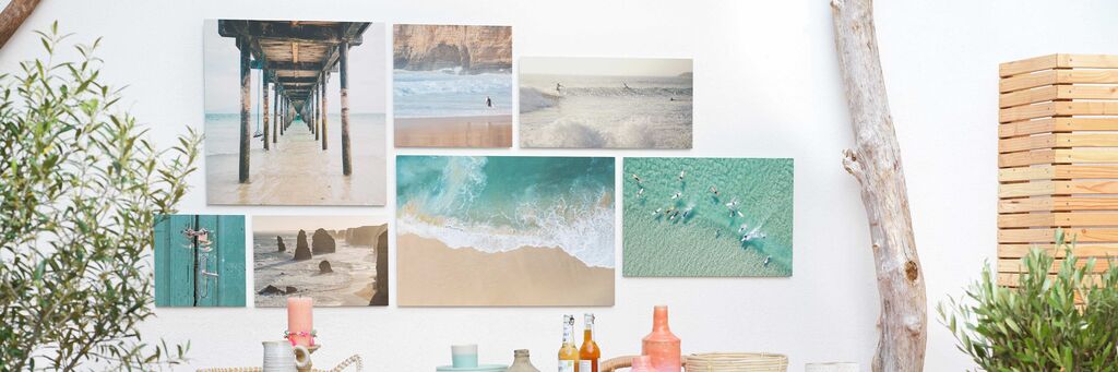 Eine kreative Anordnung von verschiedenen CEWE Wandbildern zeigt Erinnerungsmomente im Urlaub. Die Bilder zeigen Strandszenen, Aktivitäten im Wasser und eine schöne Holzstruktur.