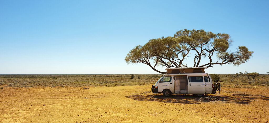 Ein gemütlicher Camper steht unter einem großen Baum in der weiten, sonnigen Wüste. Die Umgebung strahlt Ruhe und Abenteuer aus.