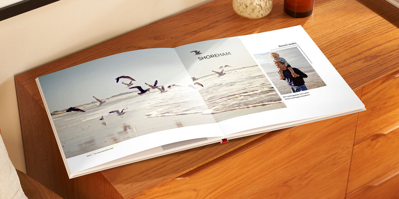 Ein geöffnetes CEWE Fotobuch auf einem Holztisch, zeigt eine Seite mit Möwen am Strand und eine persönliche Fotografie.