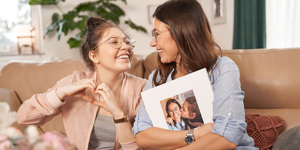 Mutter und Tochter sitzen auf dem Sofa und freuen sich über ein CEWE Fotoprodukt Geschenk, welches sie gerade auspacken.