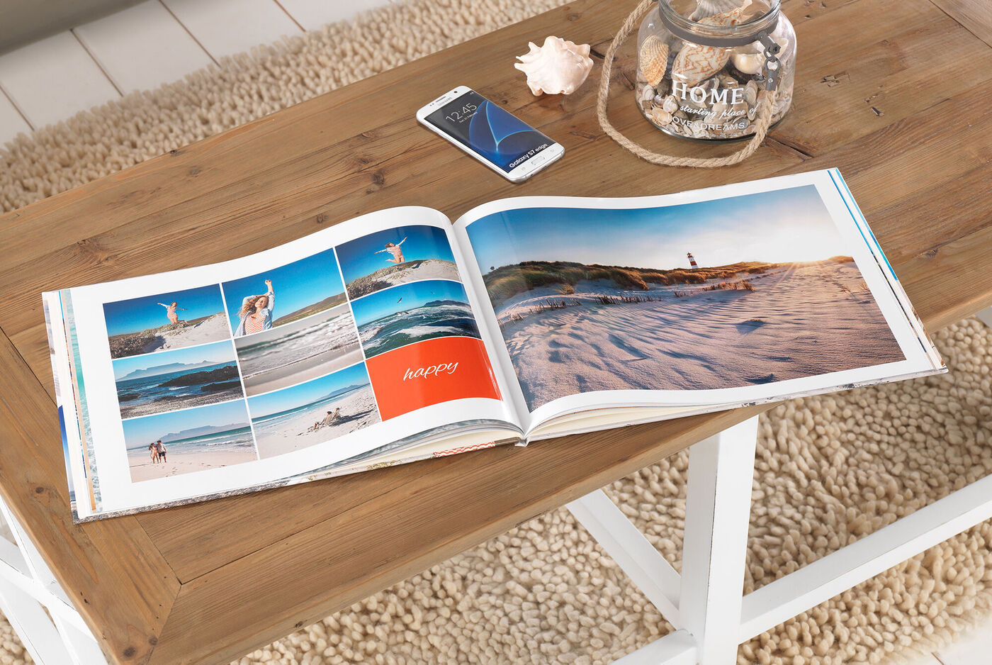 Ein auf einem Holztisch liegendes geöffnetes CEWE FOTOBUCH zeigt glückliche Strandmomente und Erinnerungen. Neben dem Buch liegt ein Handy und eine Glasvase mit Muscheln.