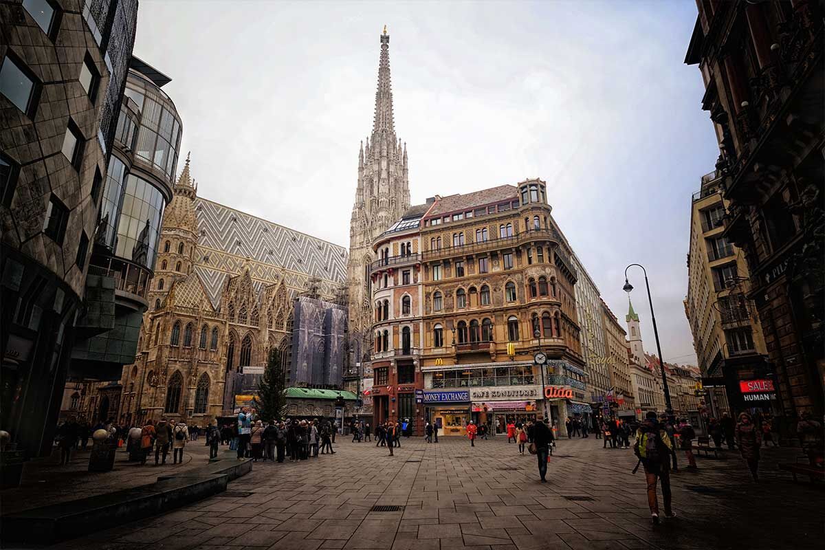 Der Stephansdom in Wien mit buntem Gebäude davor; Menschen schlendern auf dem Platz
