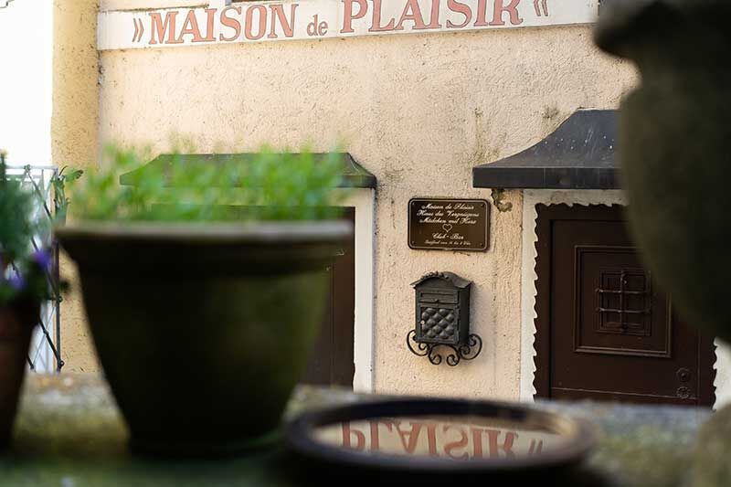 Eine charmante Fassade mit Pflanzen. Das Schild mit "MAISON de PLAISIR" sowie eine Briefkasten sind gut sichtbar.