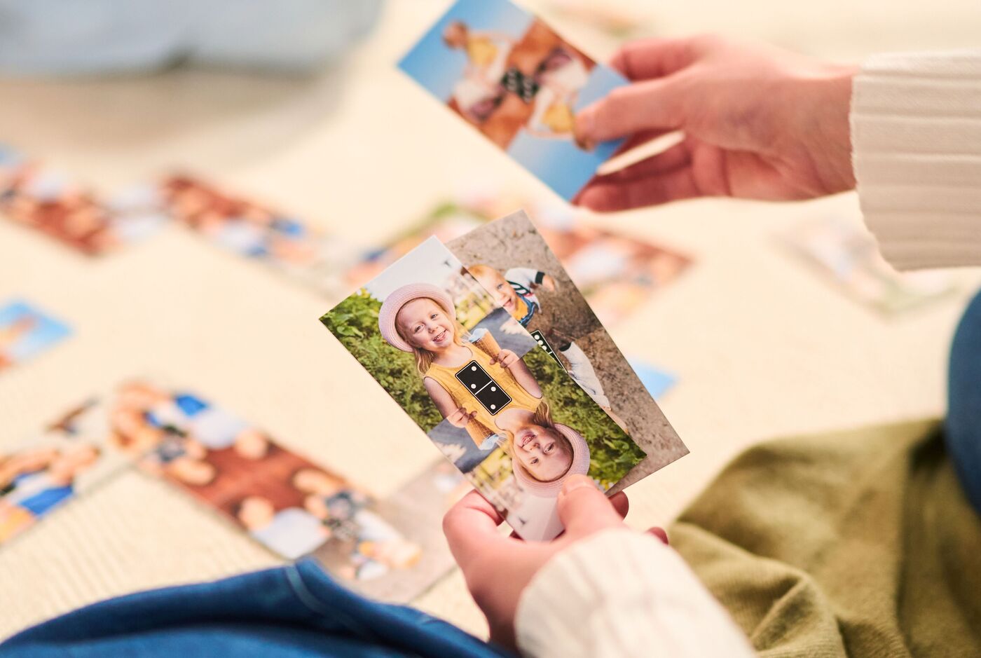 Eine Person hält zwei Fotos in den Händen und schaut auf mehrere Fotos, die auf einem Teppich liegen. Die Bilder zeigen glückliche Momente.