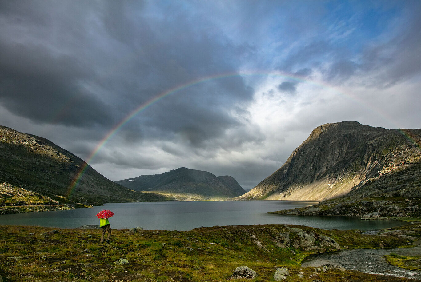 Frau mit rotem Regenschirm steht am See unter einem Regenbogen