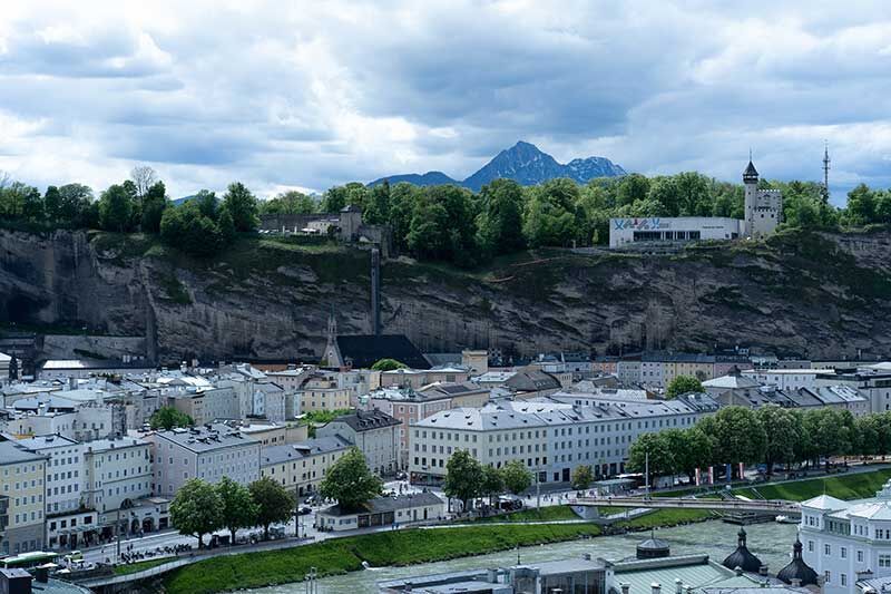 Panoramablick auf die Altstadt von Salzburg mit grüner Uferpromenade und festungsartigem Berg im Hintergrund.
