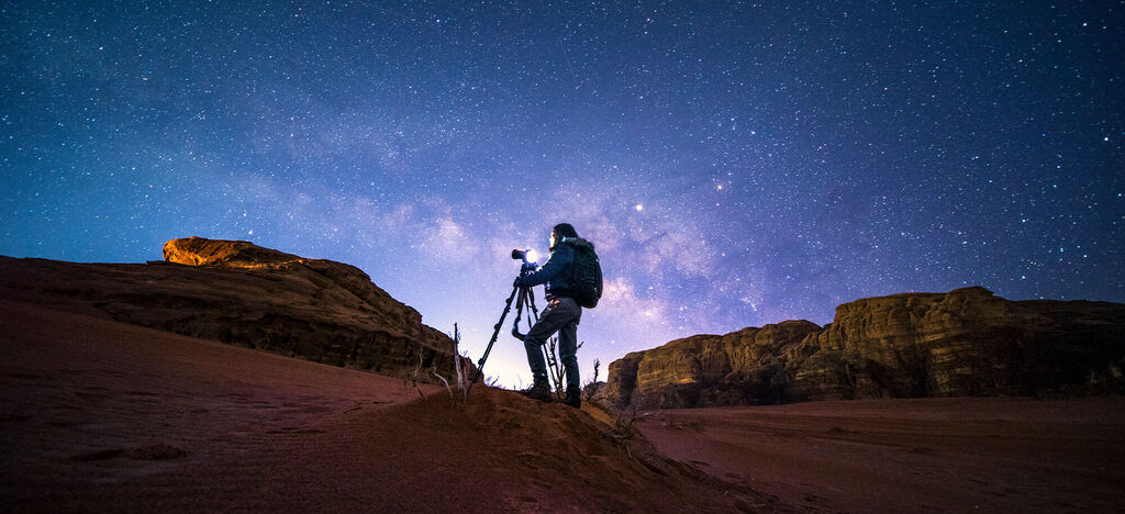 Ein Fotograf steht mit seiner Kamera in einer Wüstenlandschaft unter einem klaren Sternenhimmel.