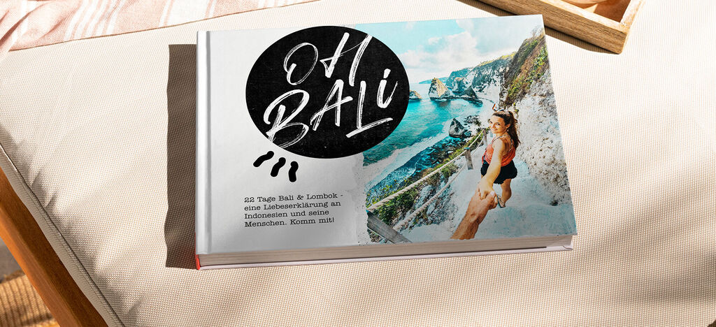 Ein CEWE FOTOBUCH mit dem Titel "Oh Bali" liegt auf einem hellen Sofa und zeigt eine farbenfrohe Landschaft mit einer Frau in einem fröhlichen Moment. Das Cover zeigt auch künstlerische Handlettering-Elemente und einen wunderschönen Blick auf die Küst