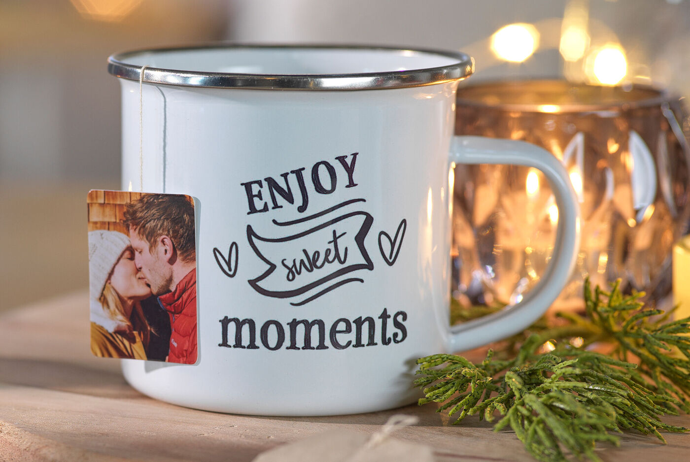 Weißer Becher mit der Aufschrift 'Enjoy sweet moments'. Ein Fotoanhänger hängt daran, daneben ist ein Teelicht und Tannenzweige.